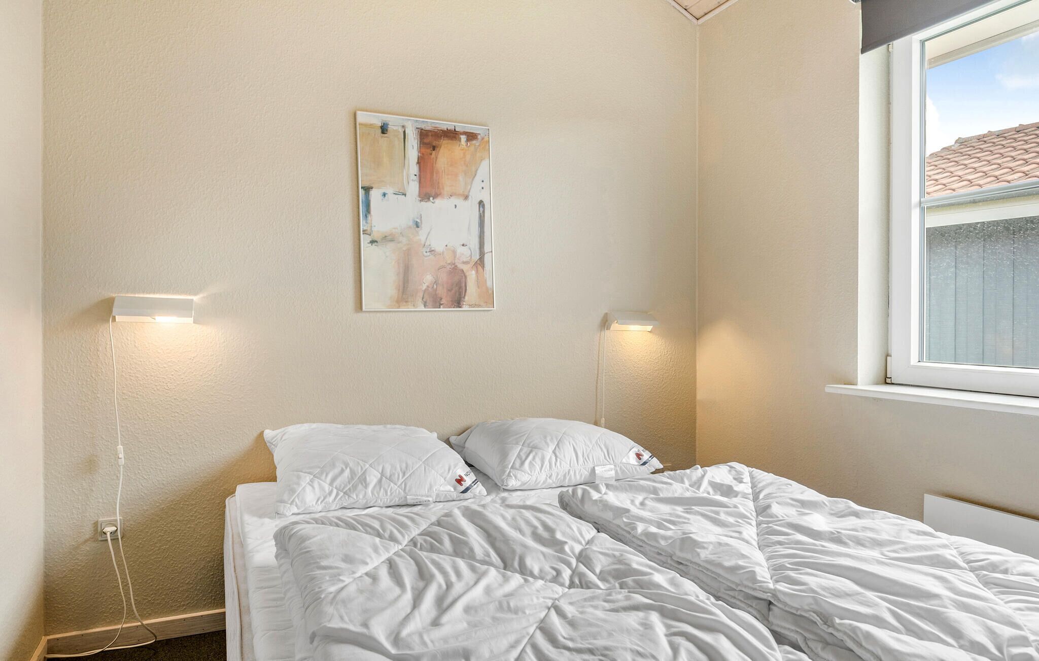 A bedroom with a bed, two bedside lamps, a painting on the wall, and a window with a view of a building.