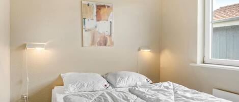 A bedroom with a bed, two bedside lamps, a painting on the wall, and a window with a view of a building.