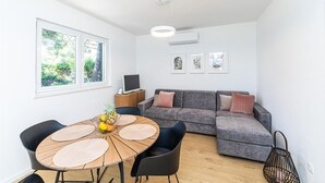 Uma sala de estar moderna com uma mesa de jantar redonda de madeira, sofá cinza e uma TV sobre um móvel de madeira.