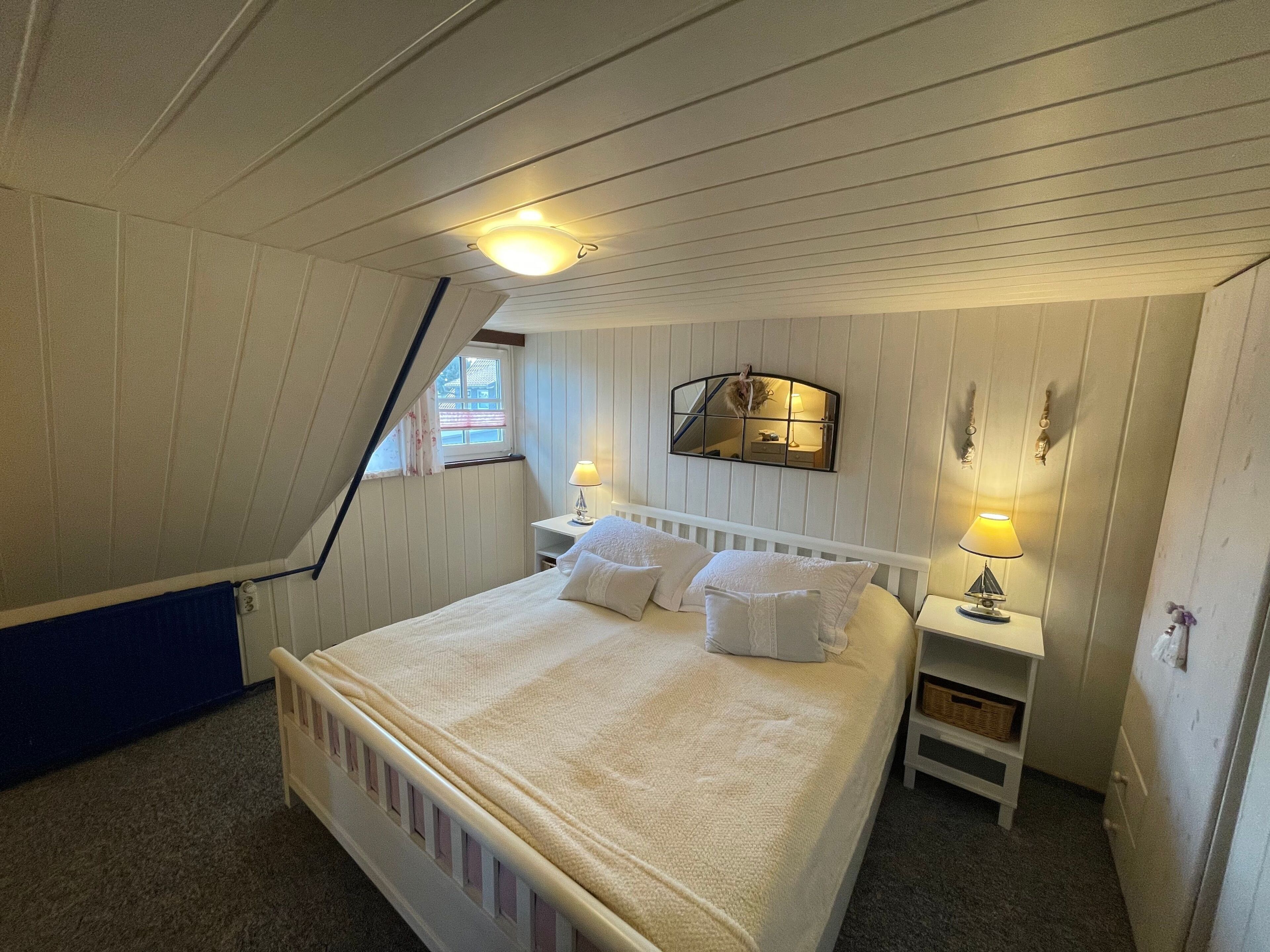 A bedroom with a sloped ceiling, a bed with white bedding, two bedside tables with lamps, a mirror above the bed, and a window with curtains.