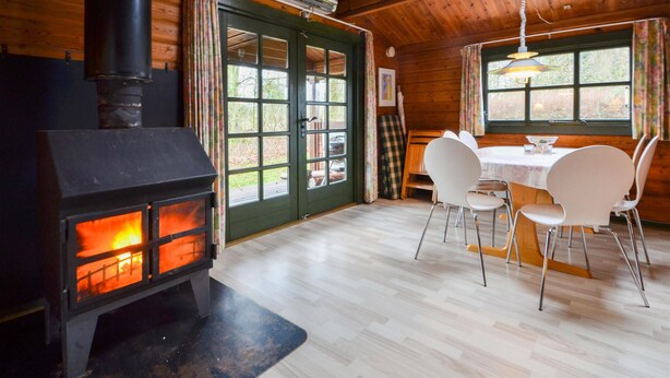 L’intérieur chaleureux d’un chalet, avec un poêle à bois, une table à manger entourée de chaises et de grandes fenêtres donnant sur le paysage extérieur.