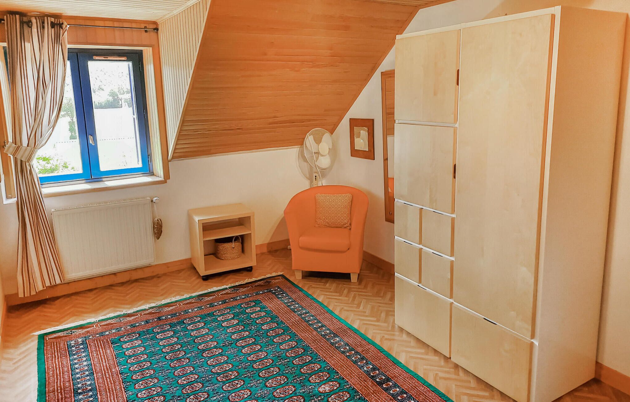 Una habitación con techo inclinado, un armario grande, una mesita con una cesta, una silla y una alfombra estampada.