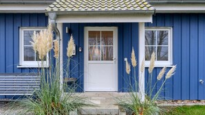 Et blåt hus med hvide døre og vinduer, en lille veranda og højt græs foran.