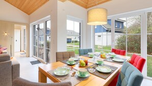 Et moderne spiseområde med et træbord dækket til morgenmad, store vinduer der giver udsigt ud, og et højt loft med synlige bjælker.