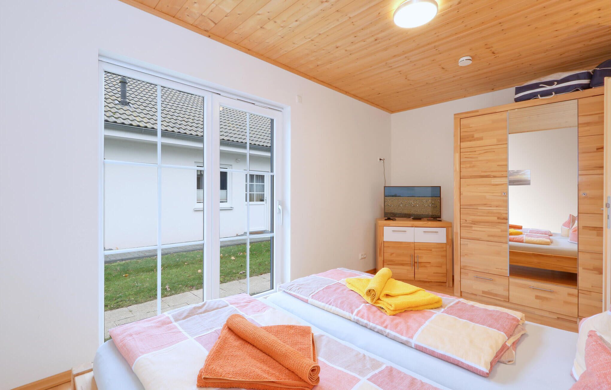 Une chambre à coucher avec un lit, une armoire en bois, un téléviseur posé sur un meuble en bois, et une grande fenêtre donnant sur un immeuble et un espace verdoyant.