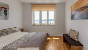 A bedroom with a large bed, a bench, a window with a view of a body of water, and a painting on the wall.