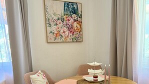Una habitación con una mesa redonda de madera, una silla con un cojín estampado, un cuadro en la pared y una ventana con cortinas.