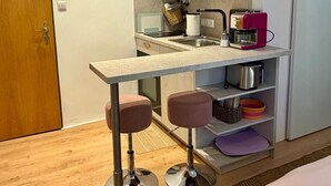 Una kitchenette compacta con un lavamanos, una cafetera y un tostador. Hay dos taburetes y un estante pequeño con vajilla.