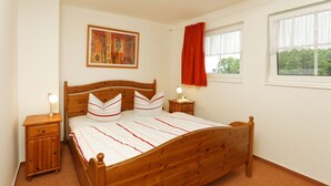 A bedroom with a wooden bed, a nightstand with a lamp, a painting on the wall, and a window with a red curtain. - Usedom tourist Koserow Hauptstr. DHH 14 - DHH 14 (Koserow)