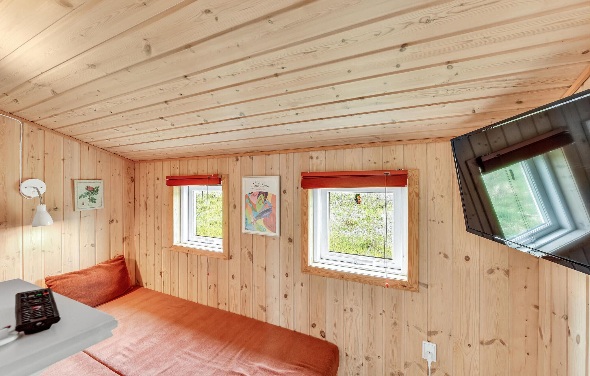 A cozy room with wooden walls, a bed, a desk with a lamp, and two windows with red frames.