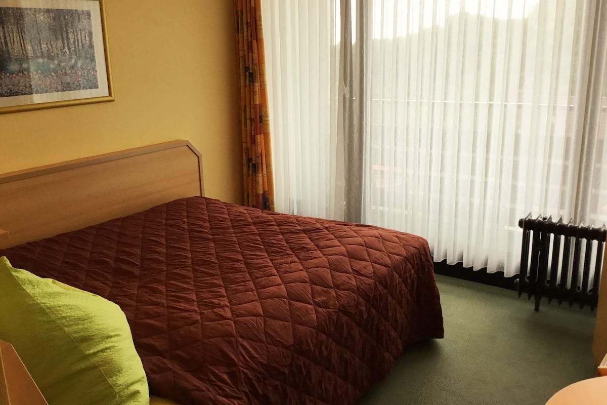 Una habitación de hotel con cama, radiador, ventana con cortinas traslúcidas y un cuadro en la pared.