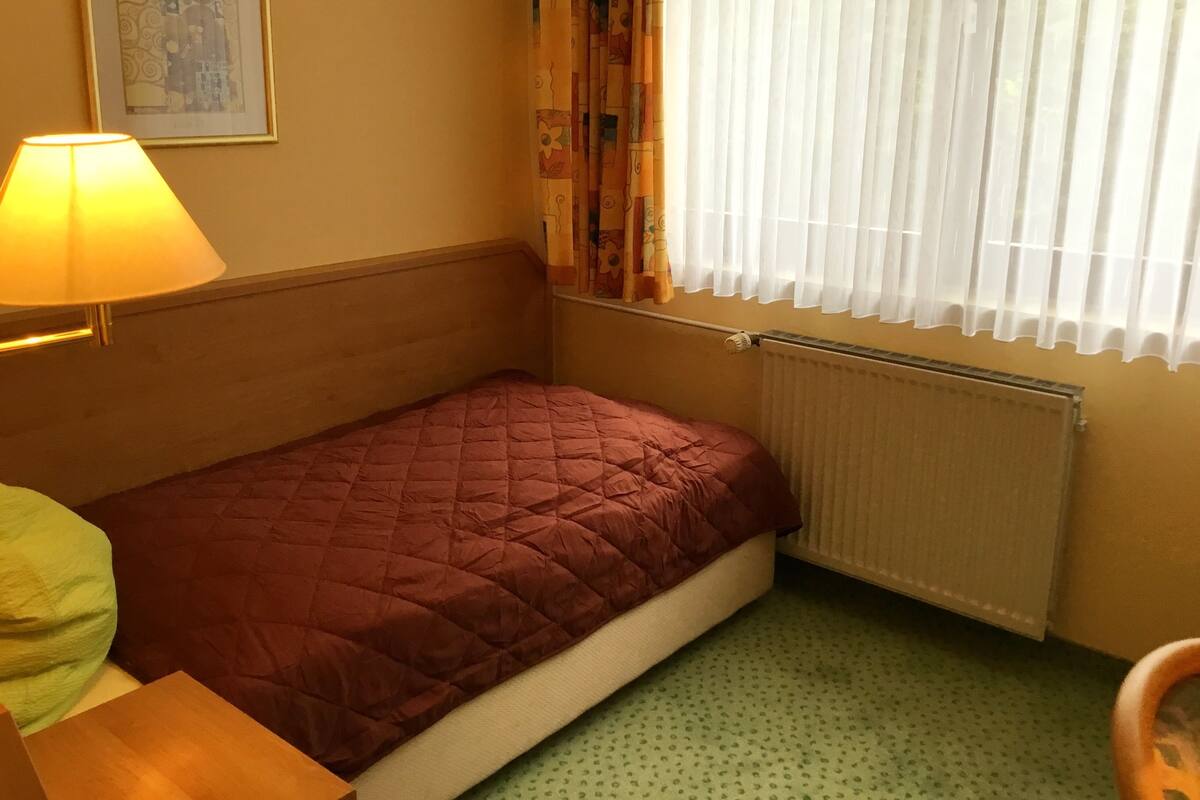 Una habitación pequeña con una cama individual, cabecera de madera, lámpara de noche, radiador y ventana con cortinas traslúcidas.
