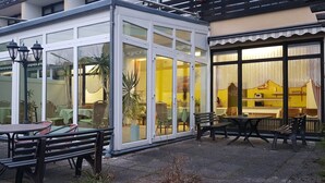 Een overdekt terras met tafels en stoelen, een glazen constructie met uitzicht op het interieur en een geel interieur zichtbaar door het glas.