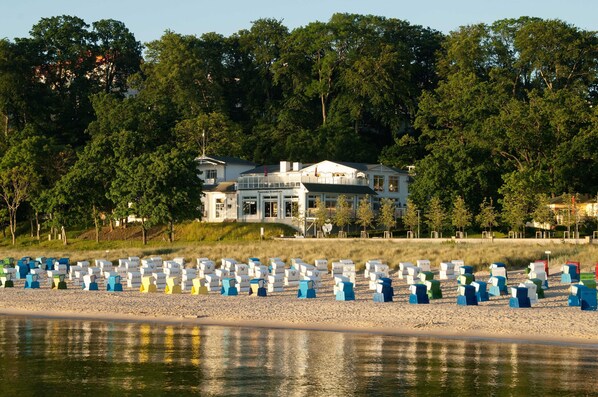 Une propriété située en bord de mer, avec une grande maison, une plage de sable fin agrémentée de chaises de plage colorées, et une végétation dense en arrière-plan.