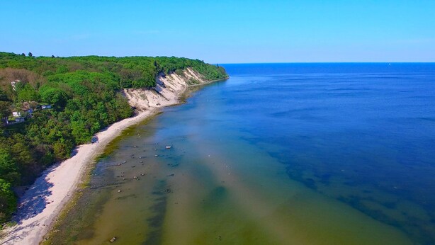 Paesaggio costiero con spiaggia sabbiosa, acqua cristallina e scogliera ricoperta di alberi.