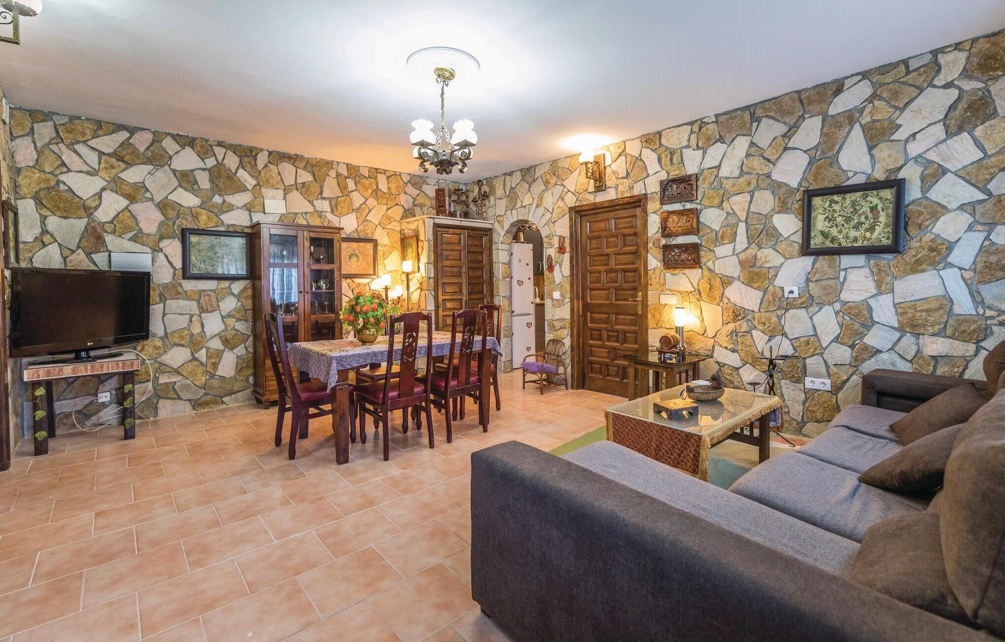 A living room with stone walls, a dining area with a table and chairs, a sofa, a TV, and a chandelier.