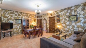 Un salon avec des murs en pierre, un coin repas avec une table et des chaises, un canapé, un téléviseur et un lustre.