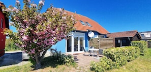 Une maison avec un toit rouge, un jardin planté d’arbres en fleurs et une terrasse aménagée avec du mobilier d’extérieur.