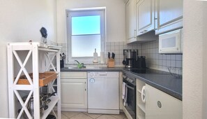 Une cuisine compacte avec des meubles blancs, un lave-vaisselle, un four à micro-ondes et une fenêtre équipée de stores.