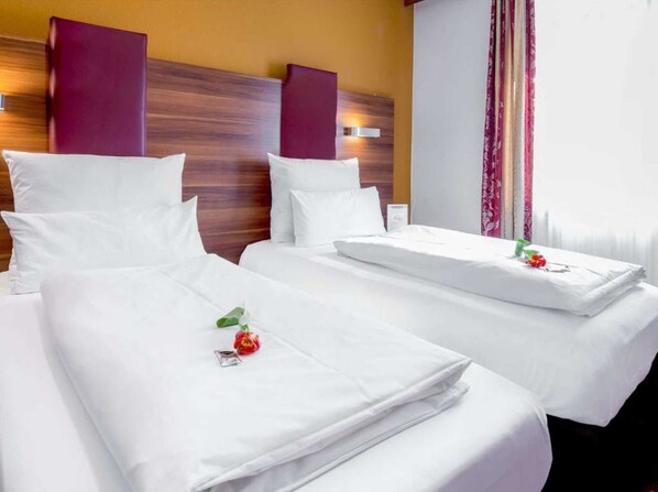 A hotel room with two beds, each with white linens and pillows, a wooden headboard, and a window with curtains.