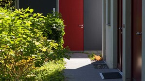 Un sendero estrecho que conduce a una puerta roja, bordeado por follaje y un muro de concreto.