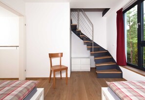 Un dormitorio con piso de madera, una silla, una cama con cubrecama a cuadros, una mesita de noche blanca, una ventana con cortina roja y una escalera con barandilla de metal.