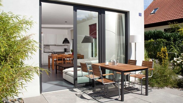 Un área de comedor exterior moderna con mesa y sillas de madera, una puerta corrediza de vidrio que da a una cocina, y un jardín con plantas y un edificio de techo rojo al fondo.