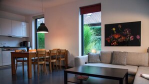 Una sala de estar moderna con sofá, mesa de centro y comedor con sillas. Hay un cuadro en la pared y una ventana con vistas a zonas verdes.