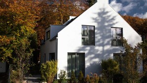 Una casa moderna blanca con un techo pronunciado, rodeada de follaje otoñal y un sendero de piedra que conduce a la entrada.