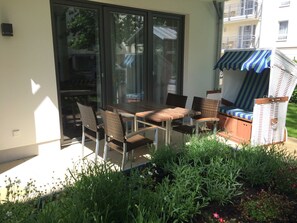 Une terrasse extérieure avec une table et des chaises, un espace salon couvert et une vue sur un immeuble d’habitation.