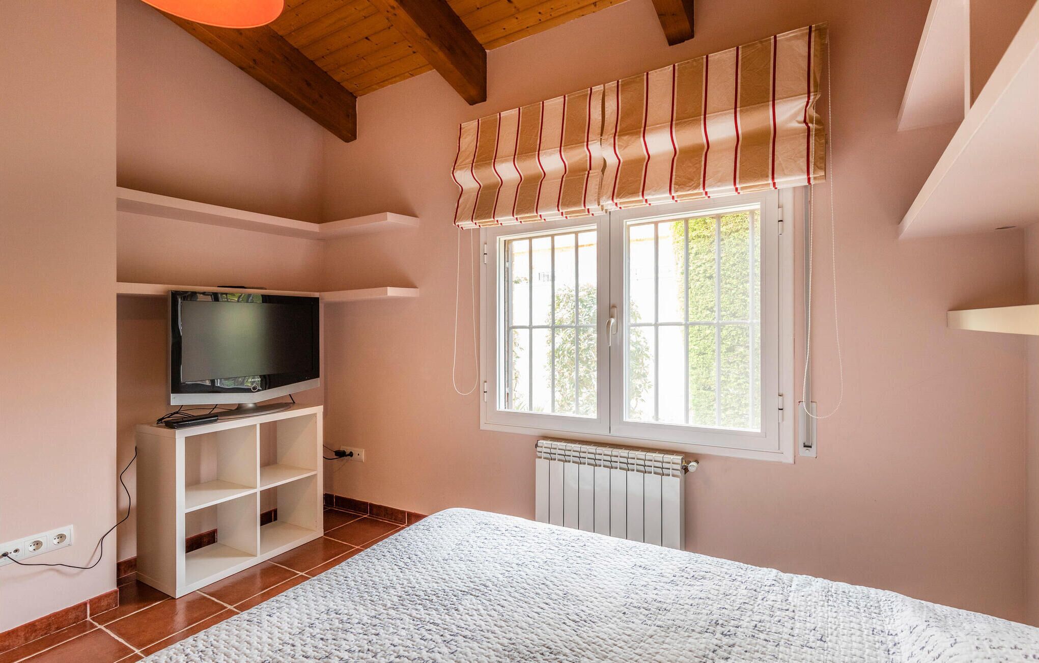 A bedroom with a television on a shelf, a window with blinds, a radiator, and a white carpet.