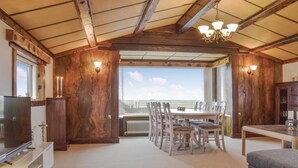 Ein geräumiger Raum mit hoher Decke, Holzbalken, einem großen Esstisch mit Stühlen, einem Fernseher und Blick durch ein großes Fenster.