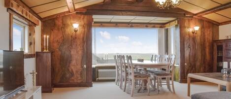 Una habitación amplia con techo alto, vigas de madera, una gran mesa de comedor con sillas, un televisor y vistas a través de un ventanal.