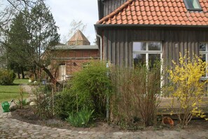 A wooden house with a tiled roof, surrounded by a garden with various plants and a stone pathway. - 2) Holiday apartment 3-room, upper floor, 90m² - Landhaus Alte Schule near (Alt Bukow)