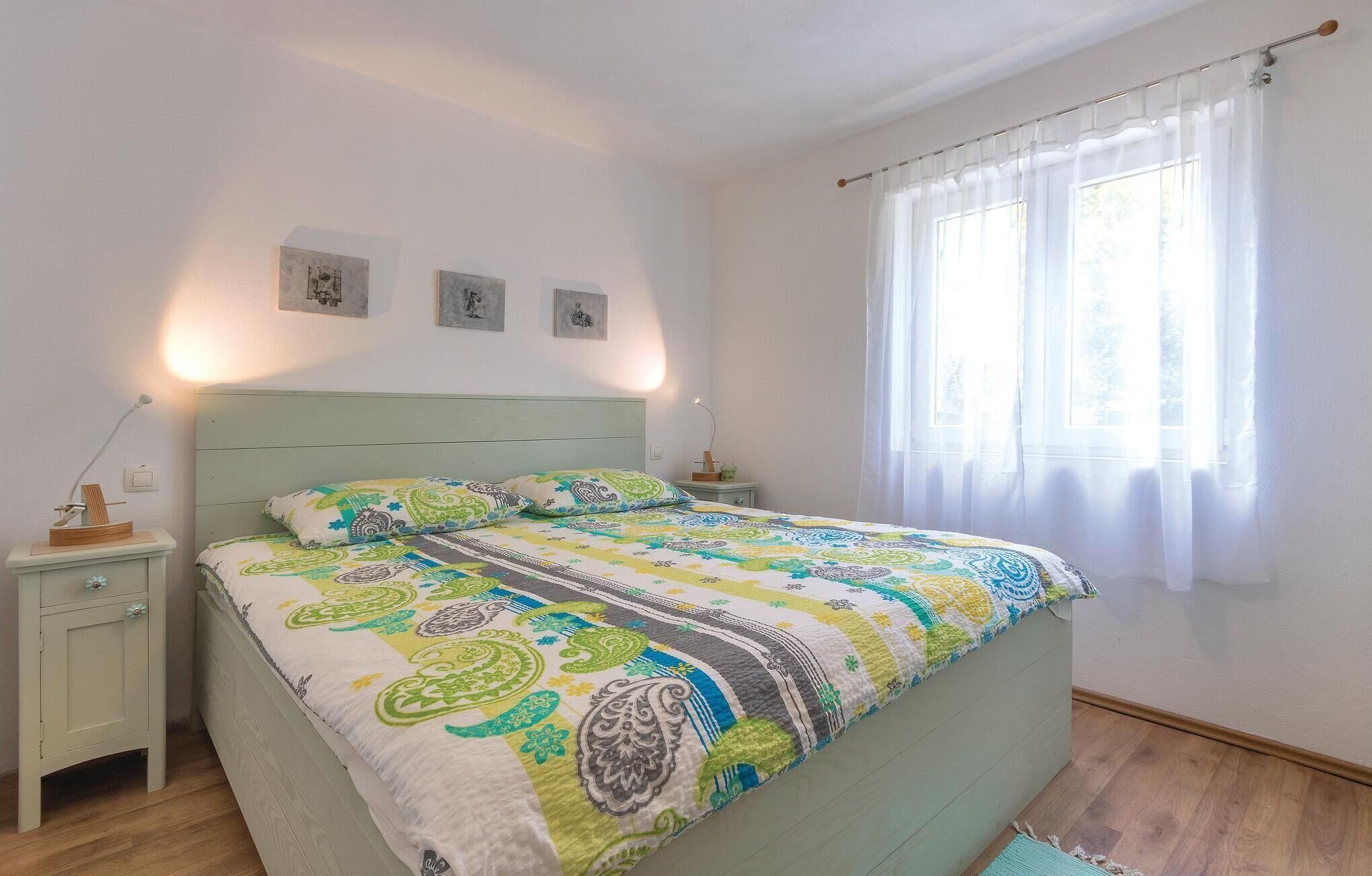 A bedroom with a bed, nightstands, a window with sheer curtains, and wall art.