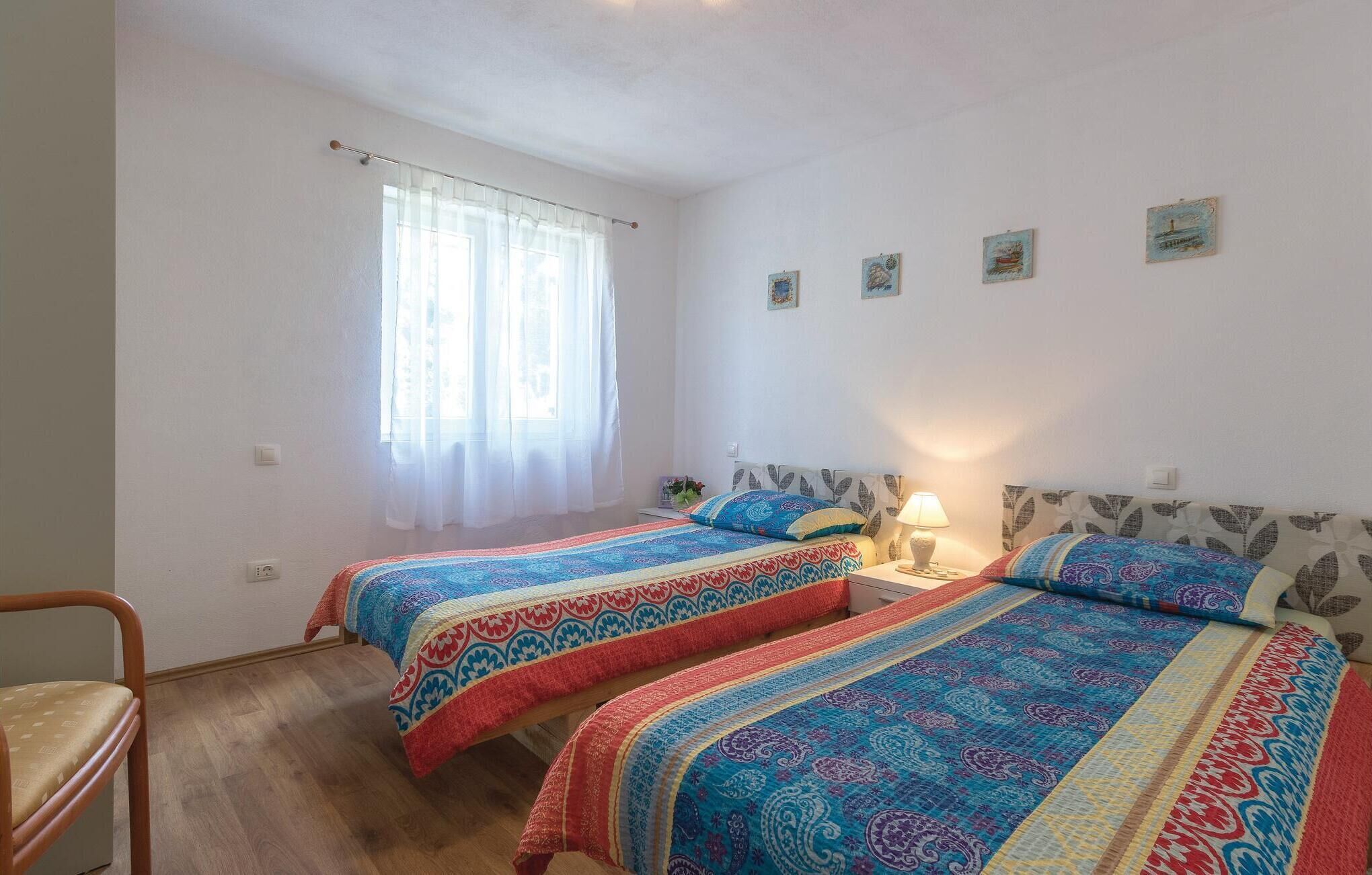 A bedroom with two beds, a window with sheer curtains, a small table with a lamp, and a chair.