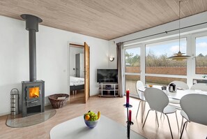 A modern living room with a wood-burning stove, a TV, and a dining area with chairs and a table. - Nice home in Rømø with sauna (Rømø)