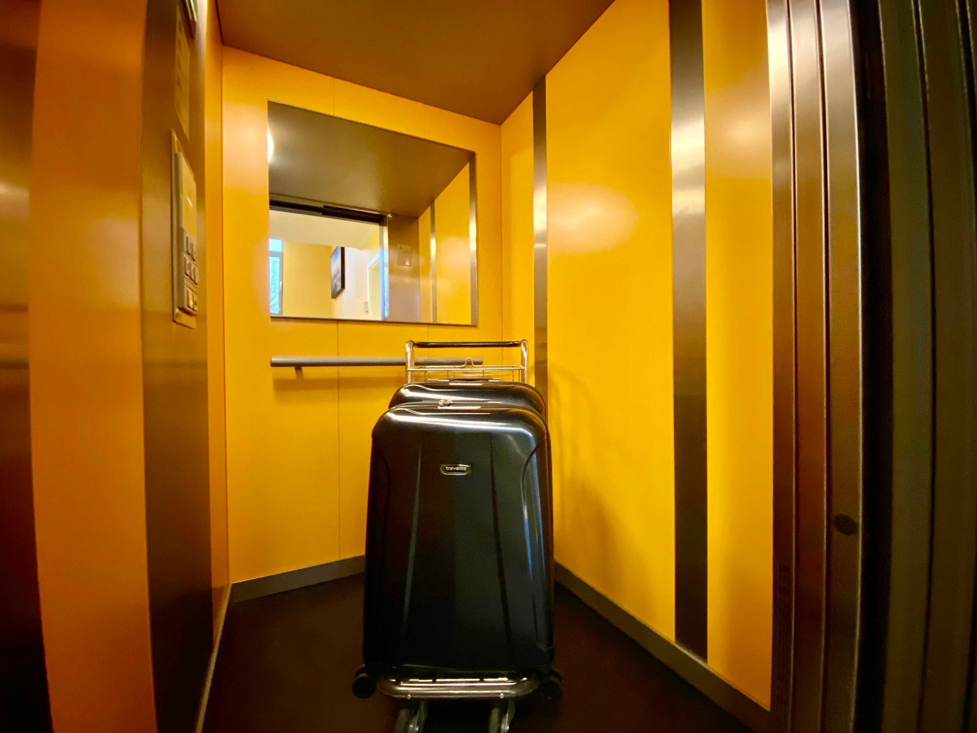 A hallway with a suitcase on a luggage cart, a mirror on the wall, and a door on the right.