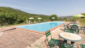 Ein Schwimmbecken mit Liegestühlen und Sonnenschirmen, umgeben von einer Steinterrasse und Grünflächen.
