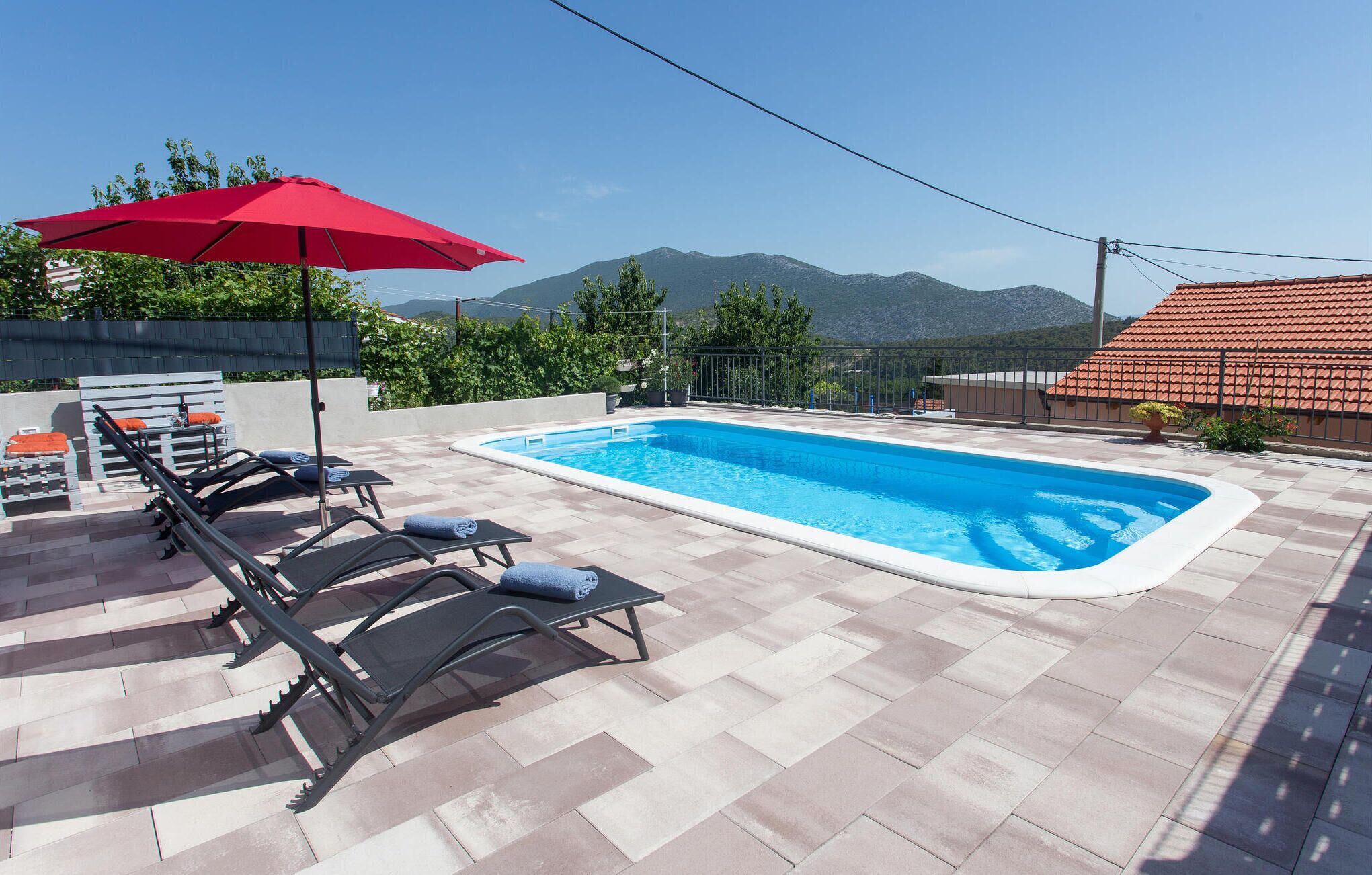 Une piscine entourée de chaises longues et agrémentée d’un parasol rouge, le tout bordé d’une terrasse carrelée et offrant une vue sur les montagnes.