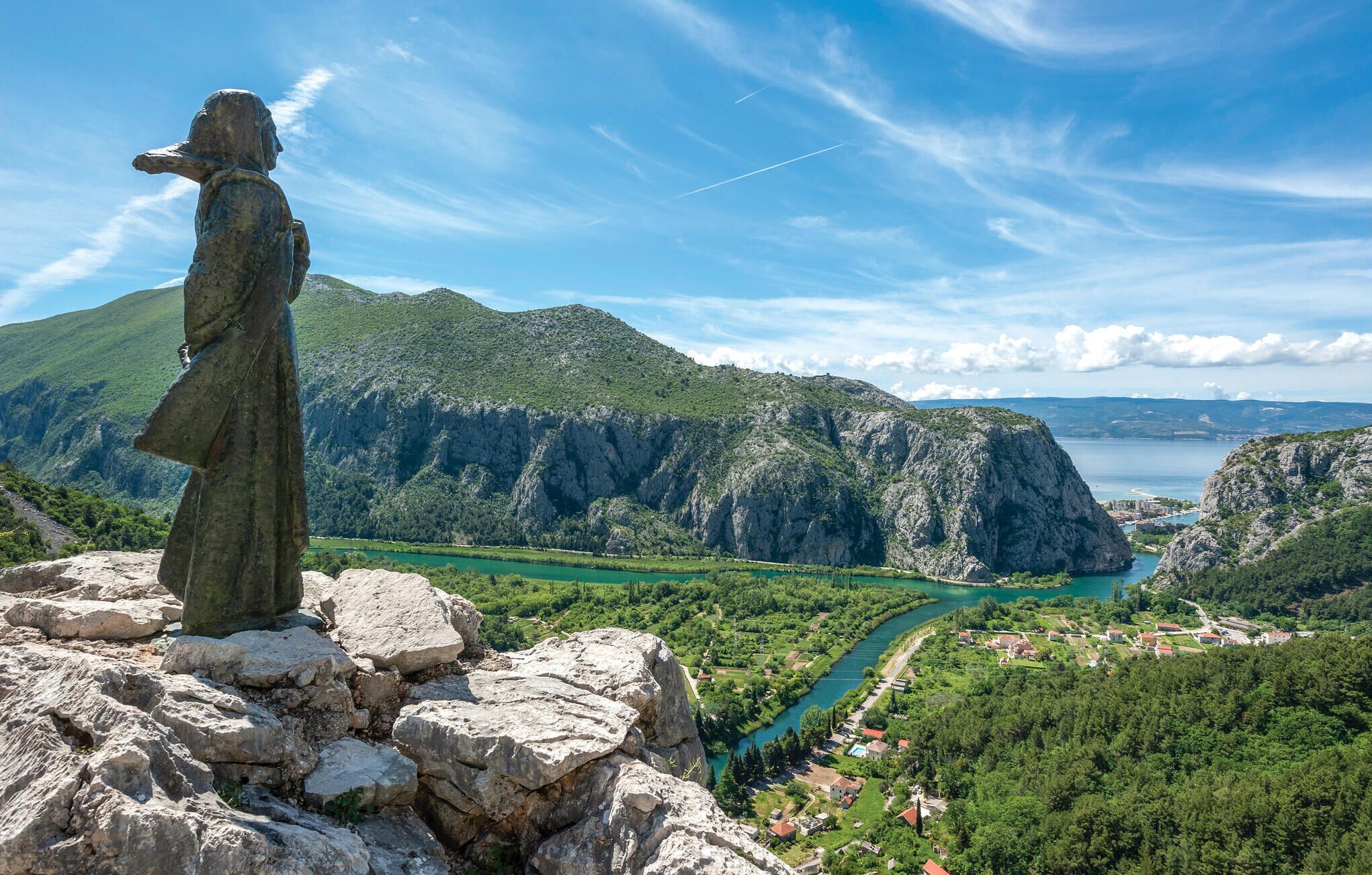 A stone statue on a rocky outcrop overlooking a valley with a river, houses, and mountains.