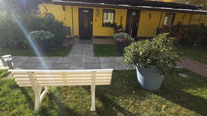 Un bâtiment jaune avec une porte noire, un banc et des plantes en pot au premier plan.