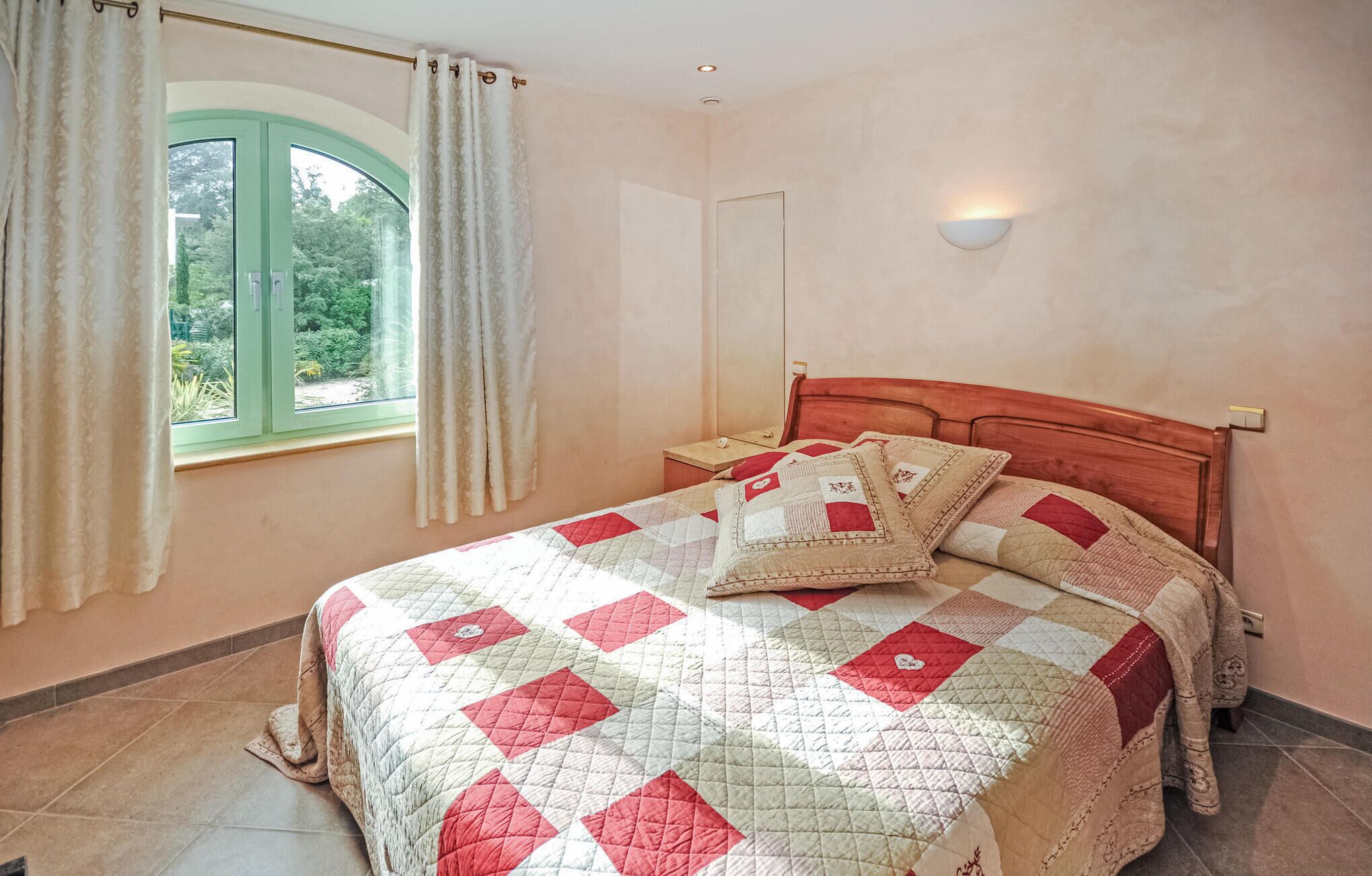A bedroom with a bed, a window with curtains, a nightstand, and a wall-mounted light.
