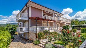 A two-story house with a balcony, a garden with flowers, and a clear blue sky.