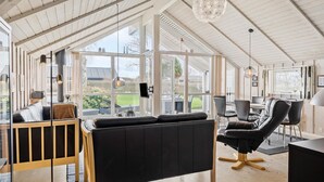Een ruime kamer met een groot raam, een zwarte leren fauteuil, een houten bankje en een eettafel met stoelen.