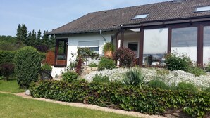 Une maison avec un toit en tuiles, un porche à l’avant et un jardin où poussent différentes plantes et fleurs.