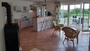 Una cocina con una isla central, un comedor con sillas de mimbre y un amplio ventanal corredero que da a un patio exterior.