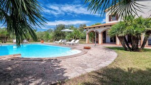 A swimming pool with lounge chairs, surrounded by palm trees and a building with a tiled roof. - Holiday home with air conditioning (Alghero)
