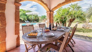 An outdoor dining area with a wooden table set for a meal, surrounded by stone arches, with a pool and greenery in the background. - Holiday home with air conditioning (Alghero)