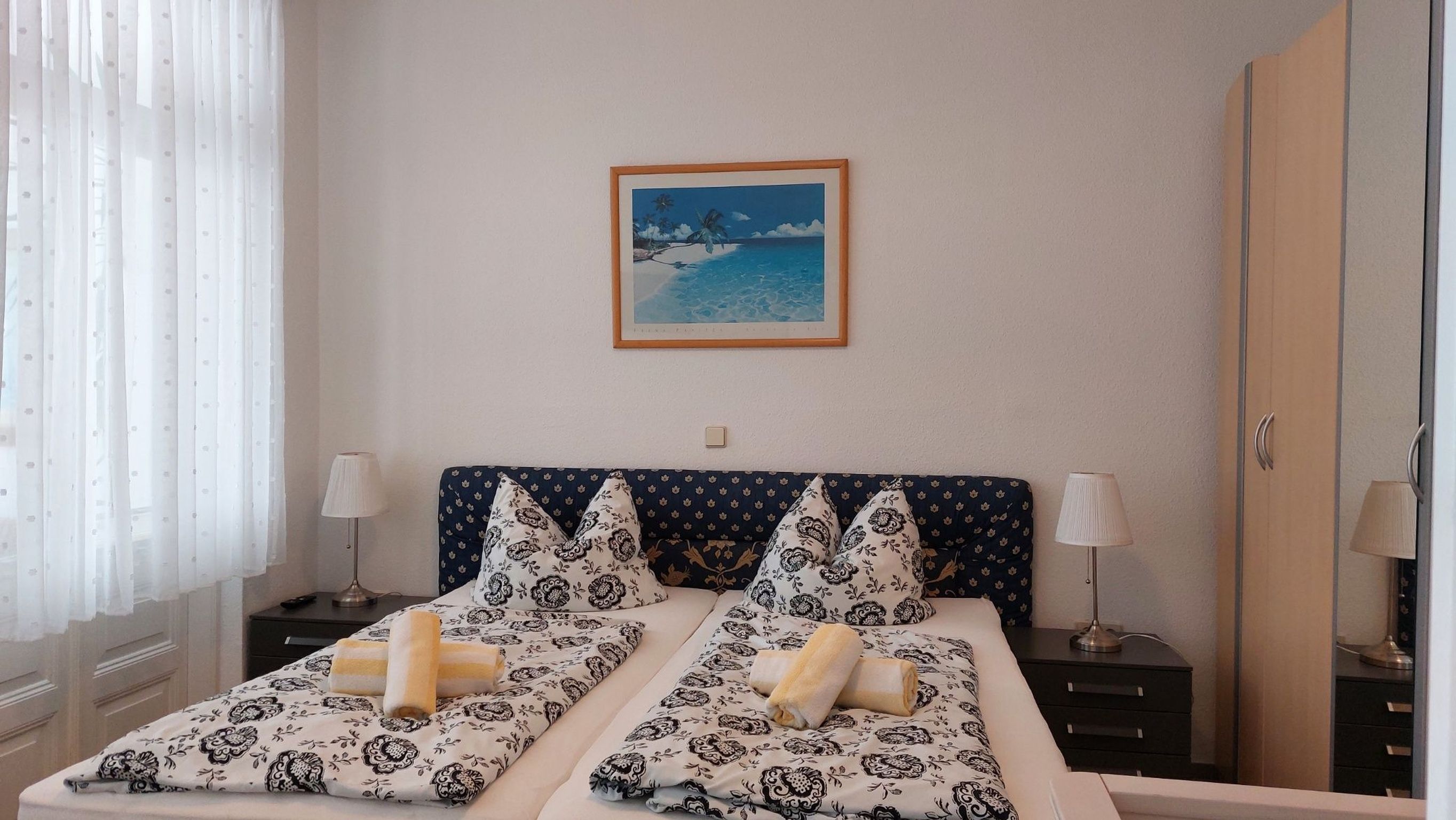 Cama de casal com roupa de cama estampada, duas mesas de cabeceira com luminárias, um quadro na parede e uma janela com cortinas leves.